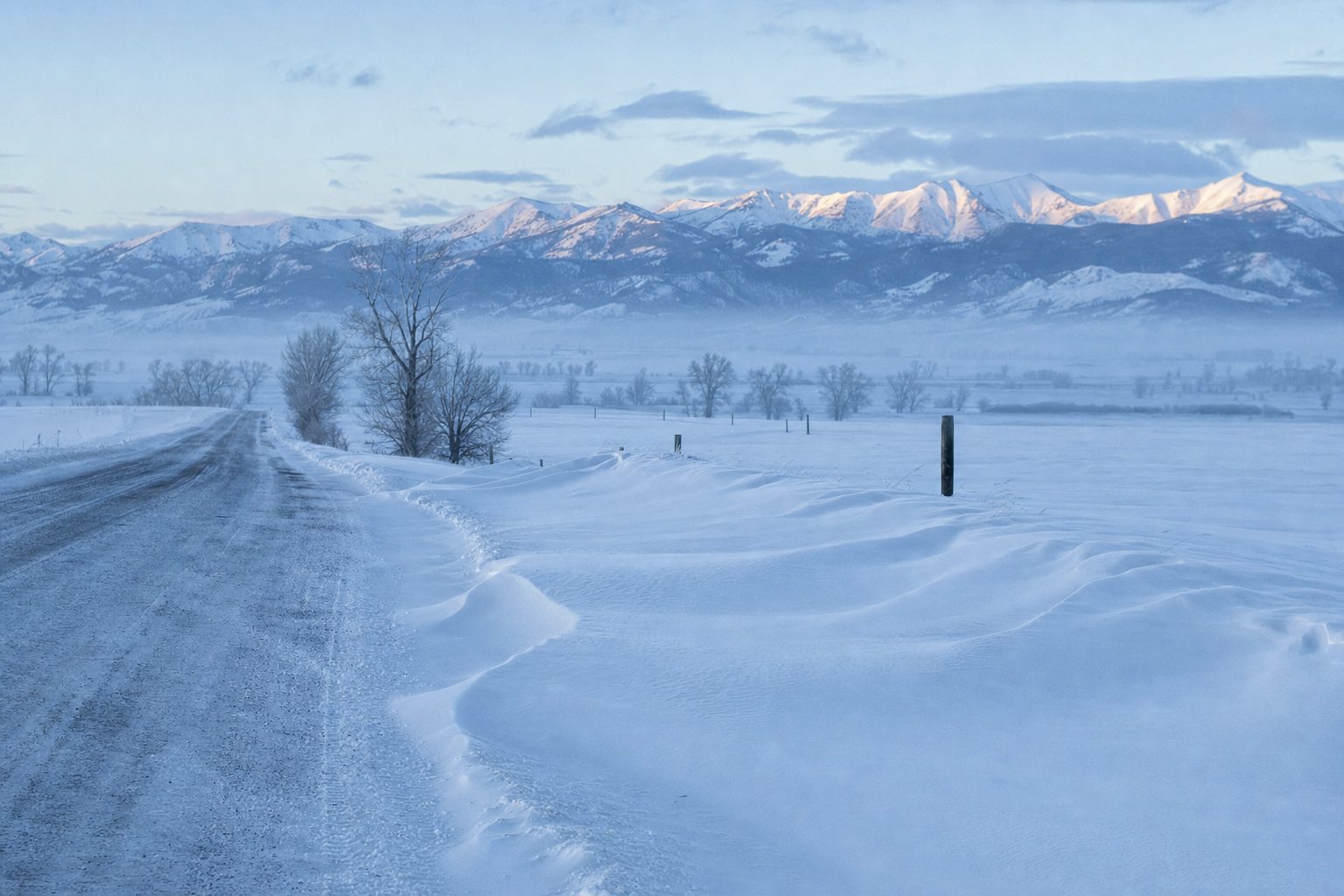 A plowed county road through deep snowdrifts in the Gallatin Valley at cold blue dusk